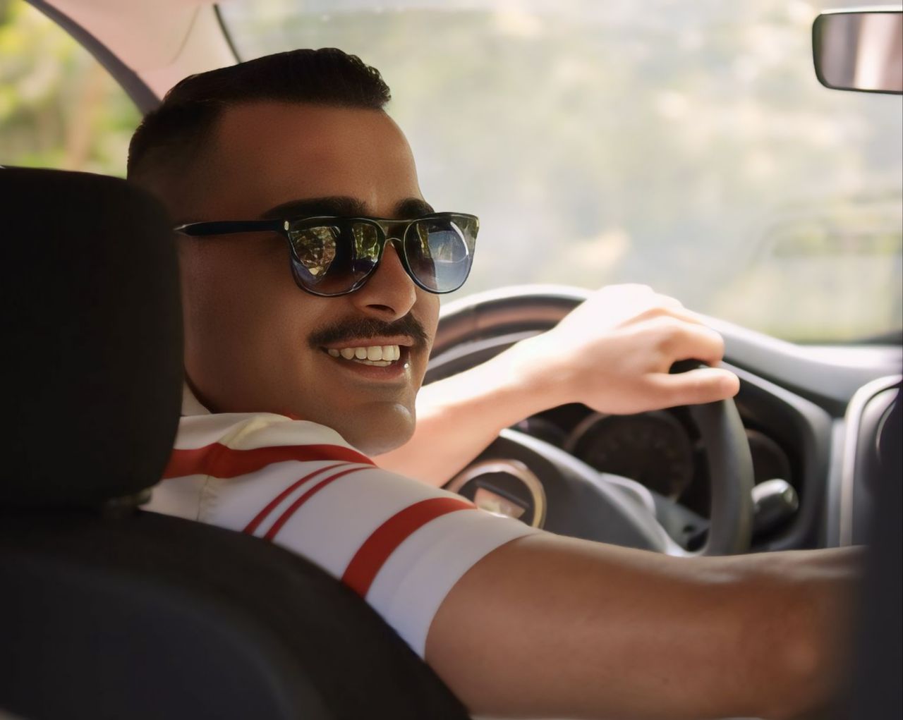 Smiling man wearing sunglasses driving a car with sunlight streaming through the window.